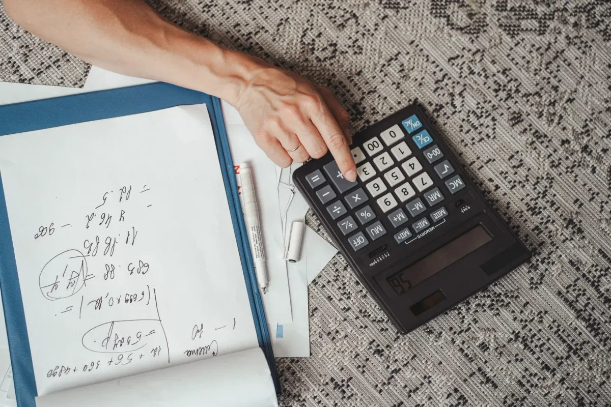 A hand using a large calculator beside handwritten notes on a desk.