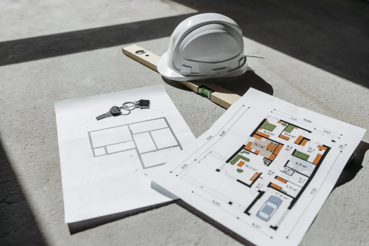 Architectural floor plans with a hard hat and keys on a sunlit floor.